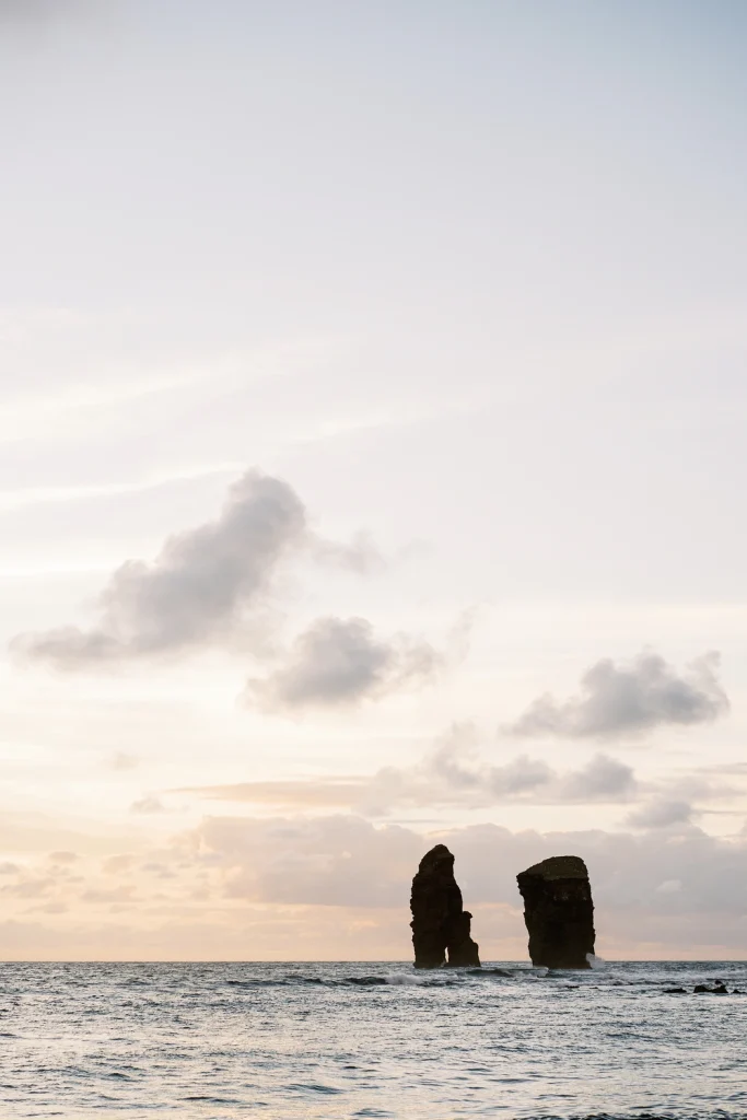 Two towering sea stacks rising from the ocean at sunset, framed by pastel-colored clouds and gentle waves in the background.