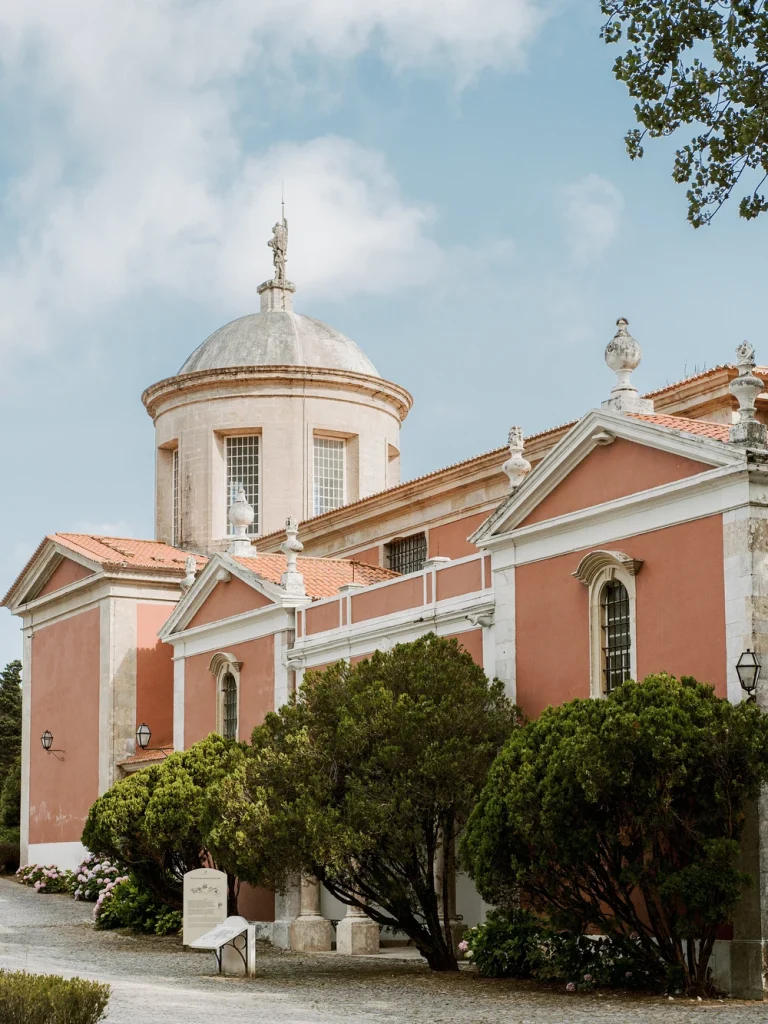 A charming pink chapel with a domed roof sits against a lush green mountain backdrop, under a clear blue sky.