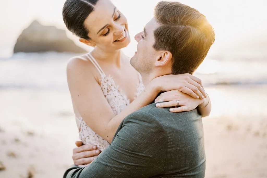 A bride wraps her arms around her groom’s shoulders as they share an intimate moment, with soft sunlight highlighting their expressions.