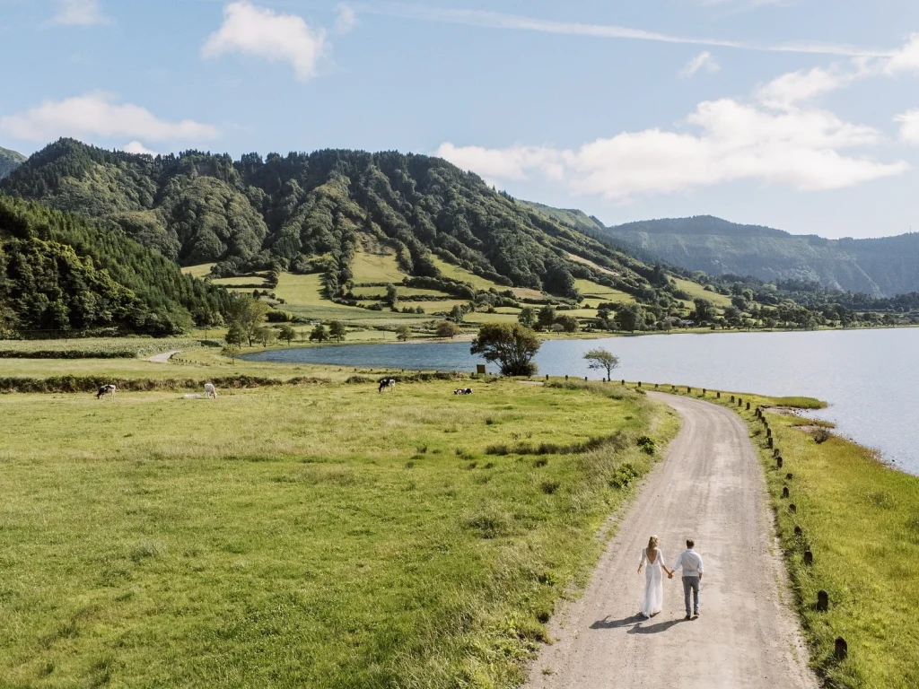 A couple walks down a scenic country road surrounded by lush green hills and a serene lake in the background.