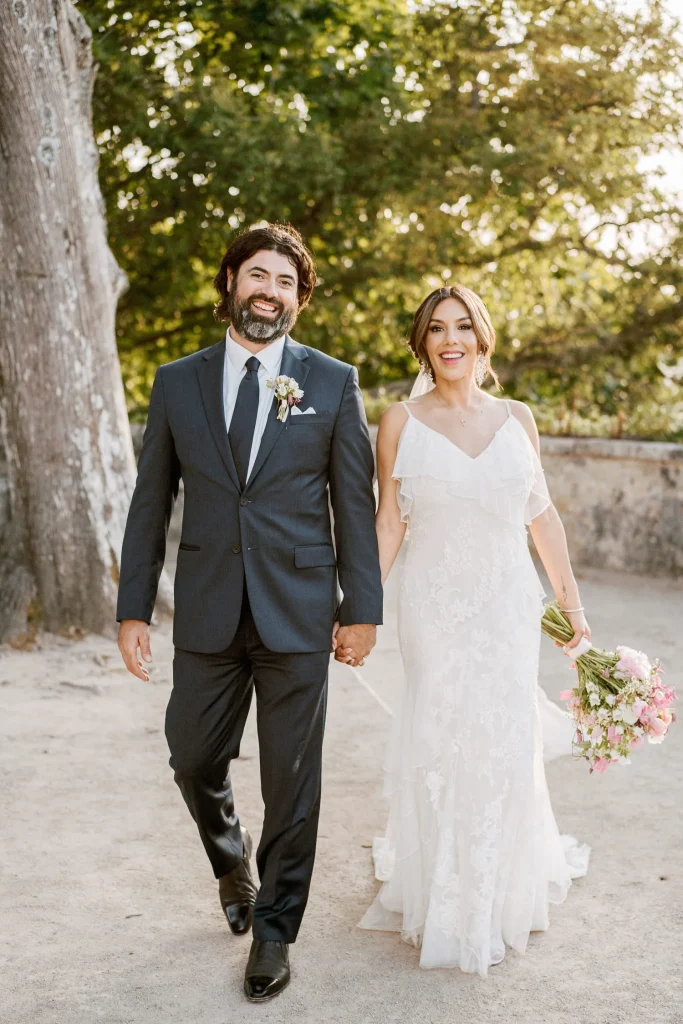 A newlywed couple walks hand in hand, smiling, with the groom in a navy suit and the bride in a flowing white dress.