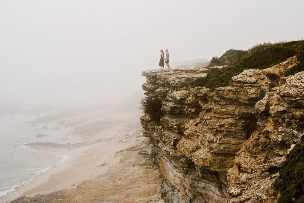 A couple stands on the edge of a dramatic cliff, overlooking the misty coastline below.