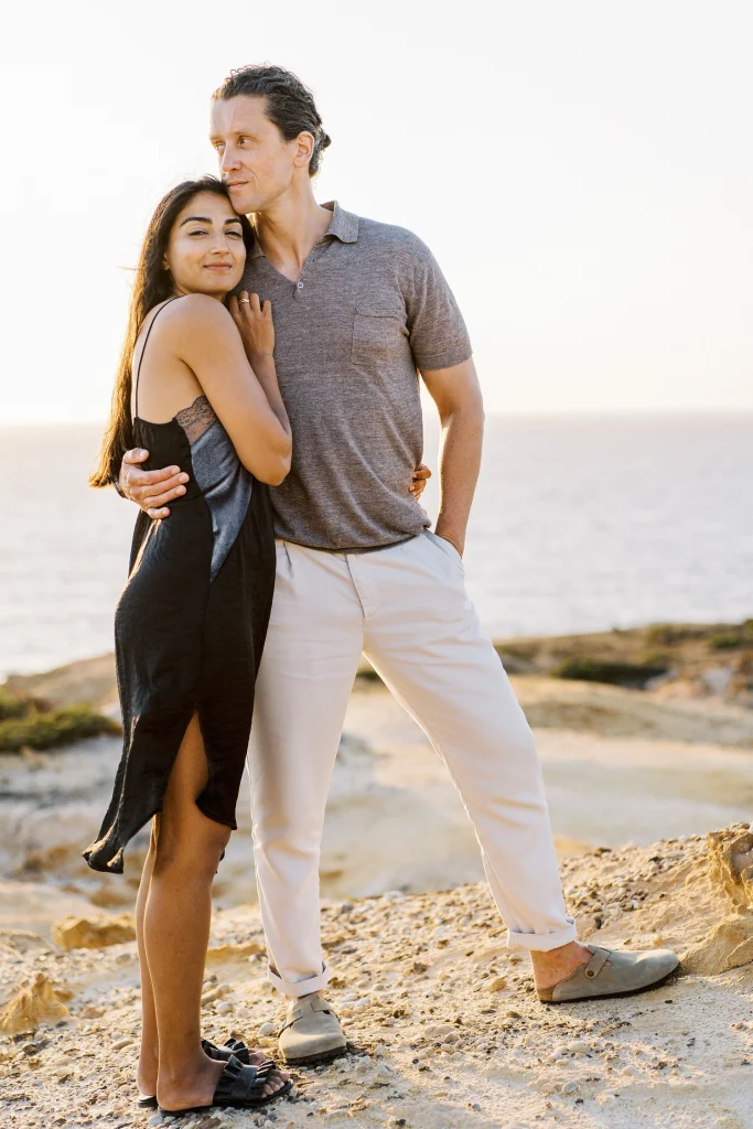 A woman wraps her arms around her partner as they stand on a scenic coastal overlook during golden hour.