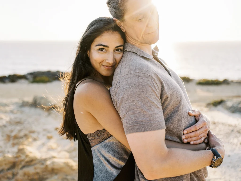 A woman smiles softly while hugging her partner from the side, with the ocean in the background.