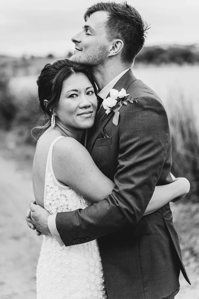 A black and white portrait of a newlywed couple embracing outdoors. The groom, in a navy suit, holds his bride close as she rests her head against his chest, looking at the camera.