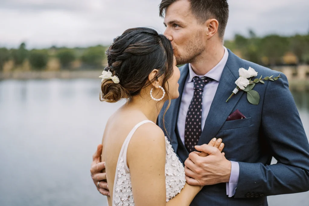 A groom in a navy suit kisses his bride's forehead as they hold hands near a serene lake. The bride, in a delicate floral wedding gown, smiles softly with flowers in her hair, capturing a quiet and intimate wedding moment.