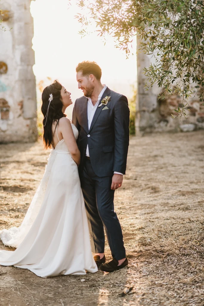 Bride and groom standing close, gazing at each other in the golden hour sunlight, surrounded by ruins and trees.