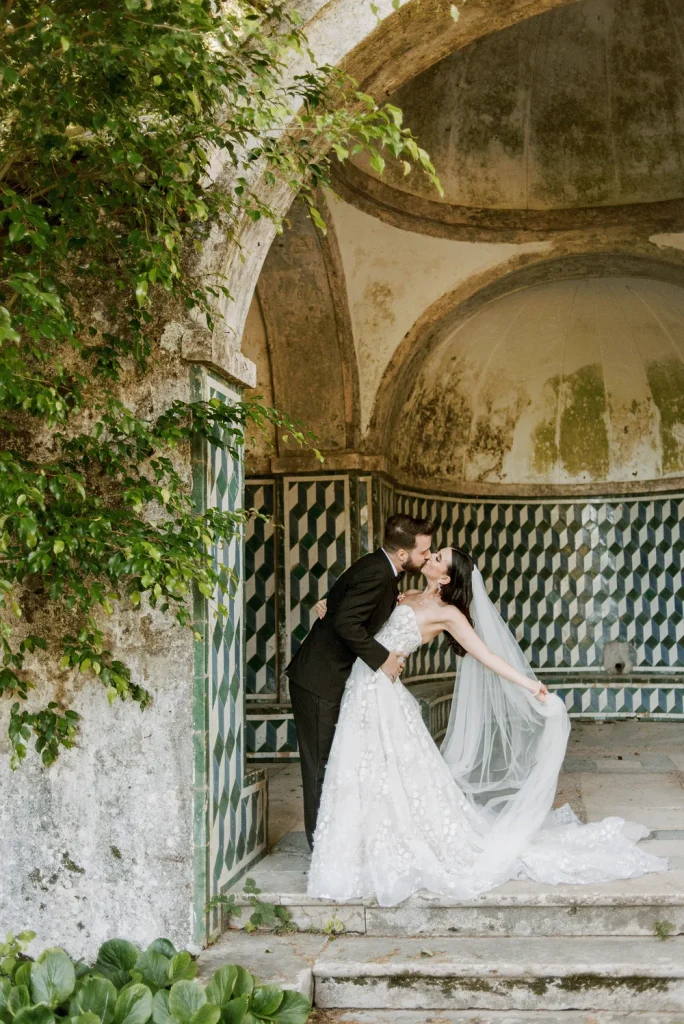 A newlywed couple shares a kiss under an elegant, weathered archway with geometric tile patterns.