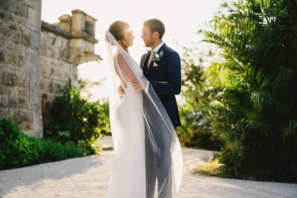 A bride and groom stand in a sunlit garden, their silhouettes framed by flowing wedding veil and historic stone architecture.