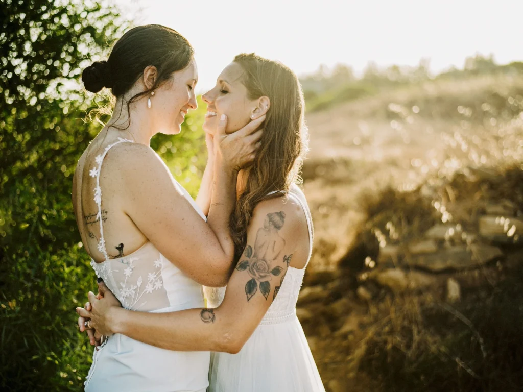 Two brides embrace warmly in a golden-lit field, gazing at each other with love.