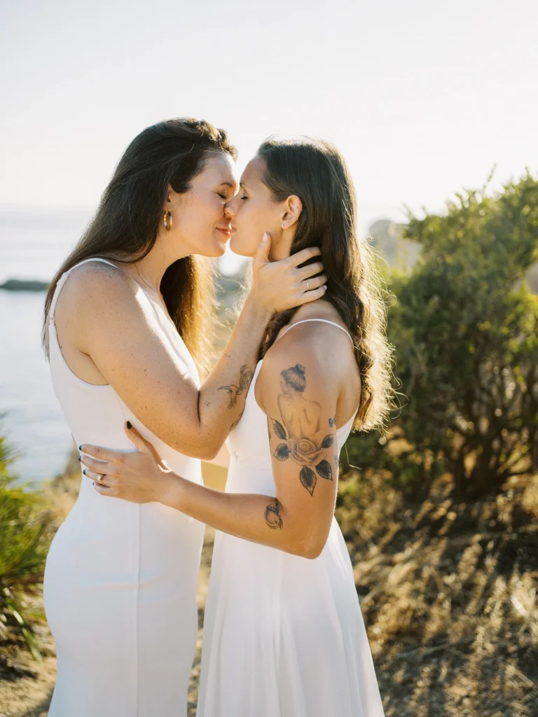 A newlywed couple shares a romantic kiss by the seaside, bathed in warm sunlight.