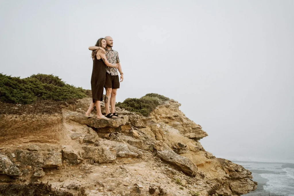 A couple embraces on the edge of a rugged cliff, looking out over the vast ocean.