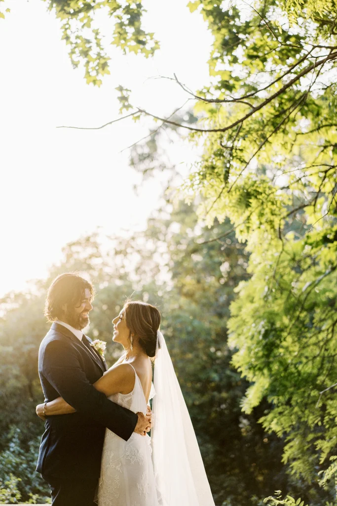 A newlywed couple gazes into each other’s eyes under golden sunlight, surrounded by lush greenery.