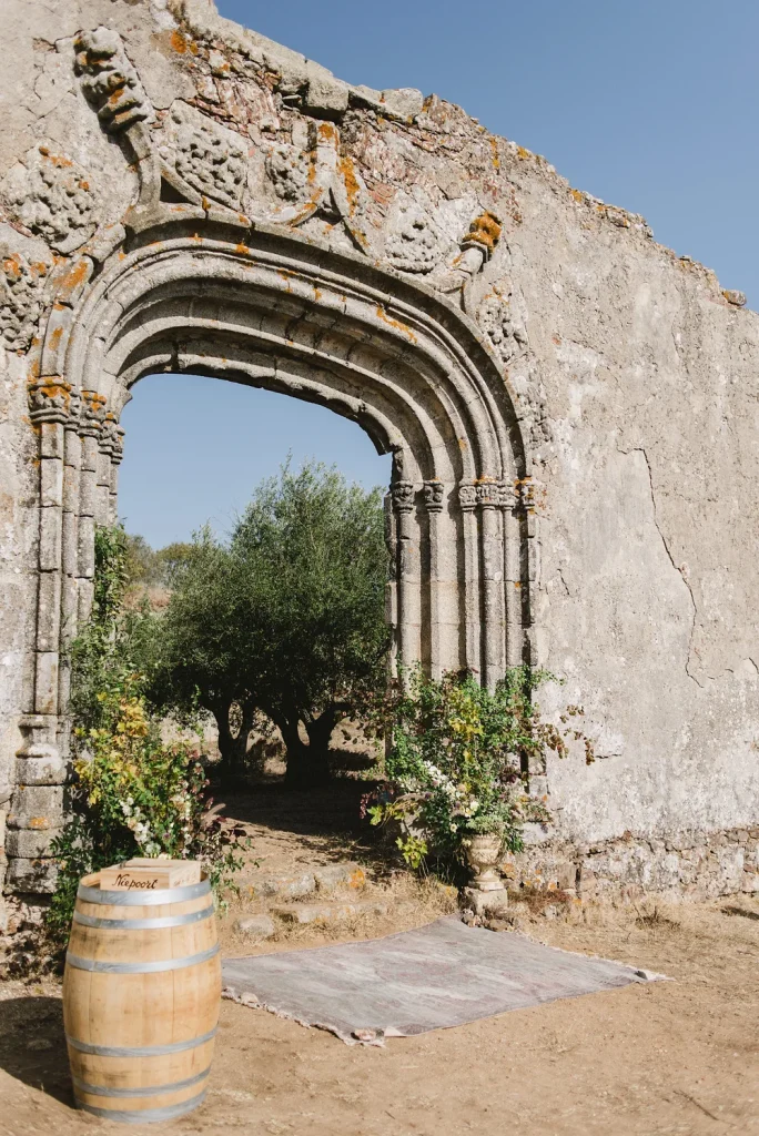 A beautifully carved stone archway from a historic ruin, leading to a sunlit grove with an olive tree.