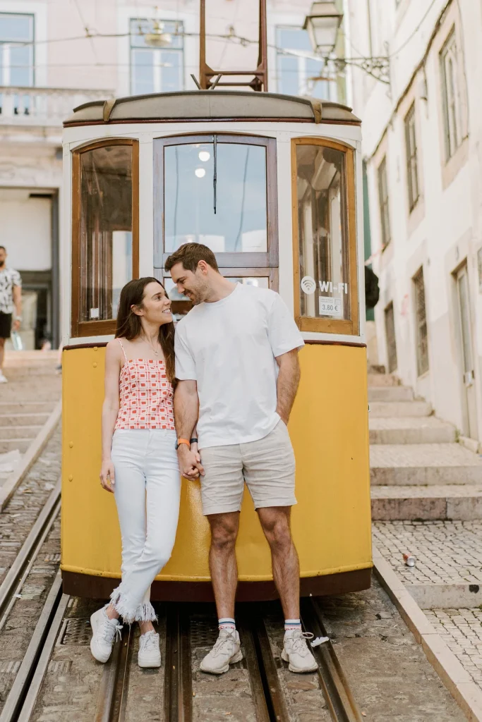 A young couple holds hands and smiles at each other in front of Lisbon’s iconic yellow tram.