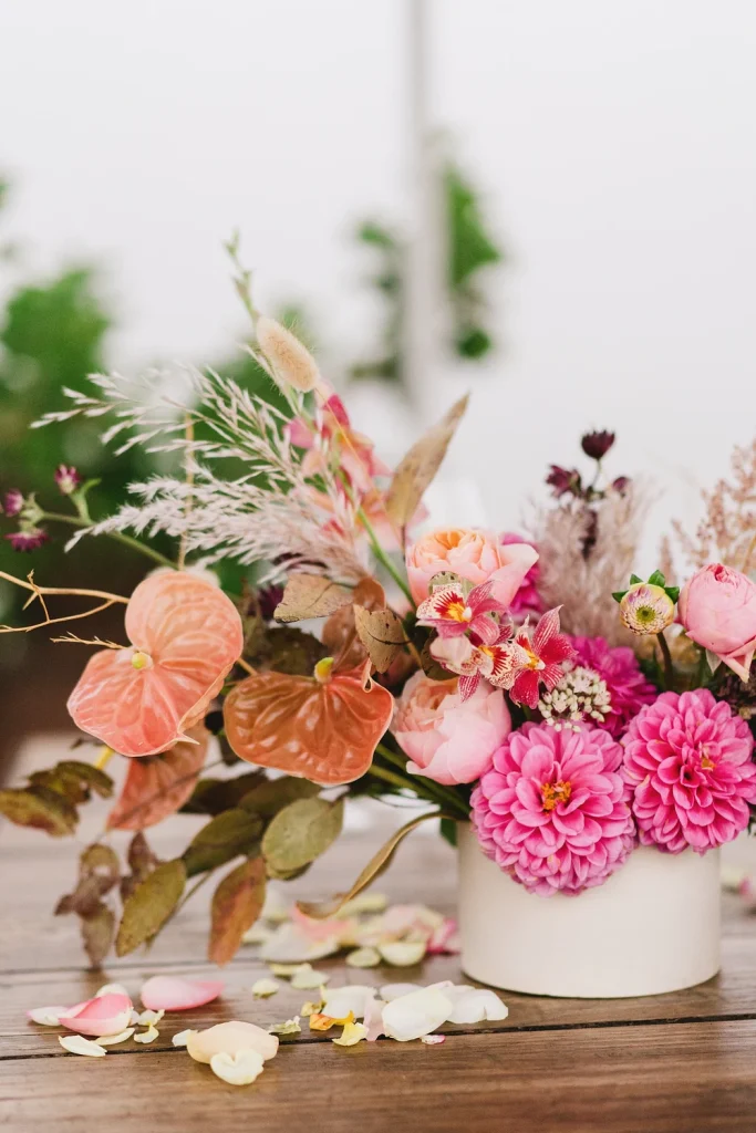 A colorful wedding flower arrangement featuring pink dahlias, roses, and exotic blooms in a white vase on a wooden table.