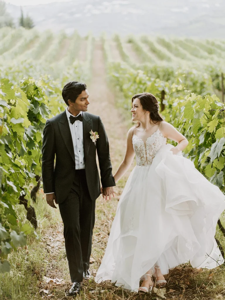 A newlywed couple walks hand in hand through lush vineyards, the groom in a classic tuxedo and the bride in a flowing white gown.