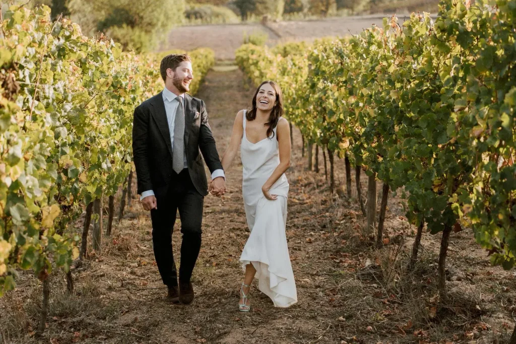 A couple strolls hand in hand through a vineyard, the bride in a simple white gown and the groom in a classic black suit.