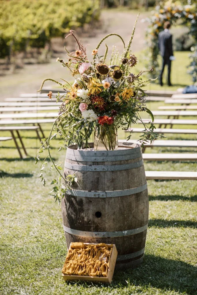 A rustic floral arrangement in warm autumn colors sits atop a wooden wine barrel at an outdoor vineyard wedding ceremony, with wooden benches arranged in the background.