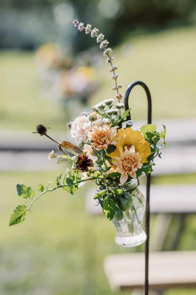 A delicate floral arrangement of peach and yellow flowers in a glass jar hangs from a shepherd’s hook along the wedding aisle, with wooden benches in the blurred background.