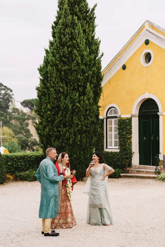 A bride in traditional Indian wedding attire shares a lighthearted moment with guests in the courtyard of Quinta de Sant’Ana, Portugal. The charming yellow-washed building and towering cypress trees add to the romantic setting.