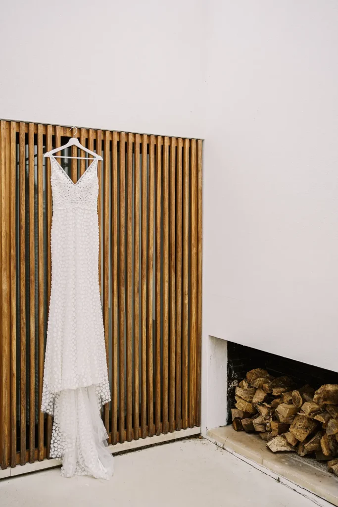 A modern white wedding dress with delicate floral details hanging on a wooden hanger against a stylish wooden slatted wall.
