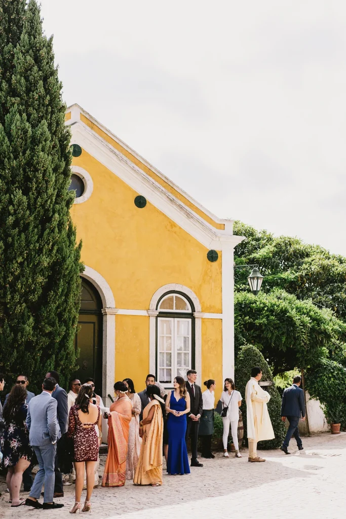 Wedding guests dressed in colorful and elegant attire gather outside a charming yellow building at Quinta de Sant’Ana, Portugal. The scene is filled with anticipation and lively conversations as they prepare for the celebration.