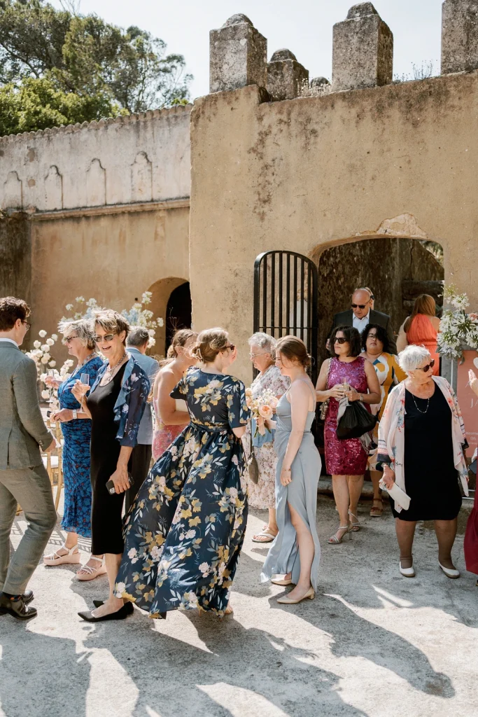 Wedding guests in elegant attire mingling outside a historic venue under the sun.