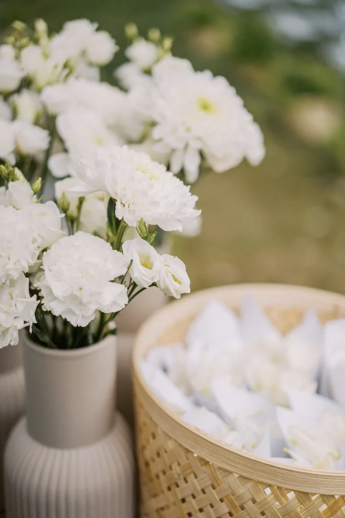 A close-up of elegant white wedding flowers in a ceramic vase, alongside a woven basket filled with white petals in delicate paper cones.