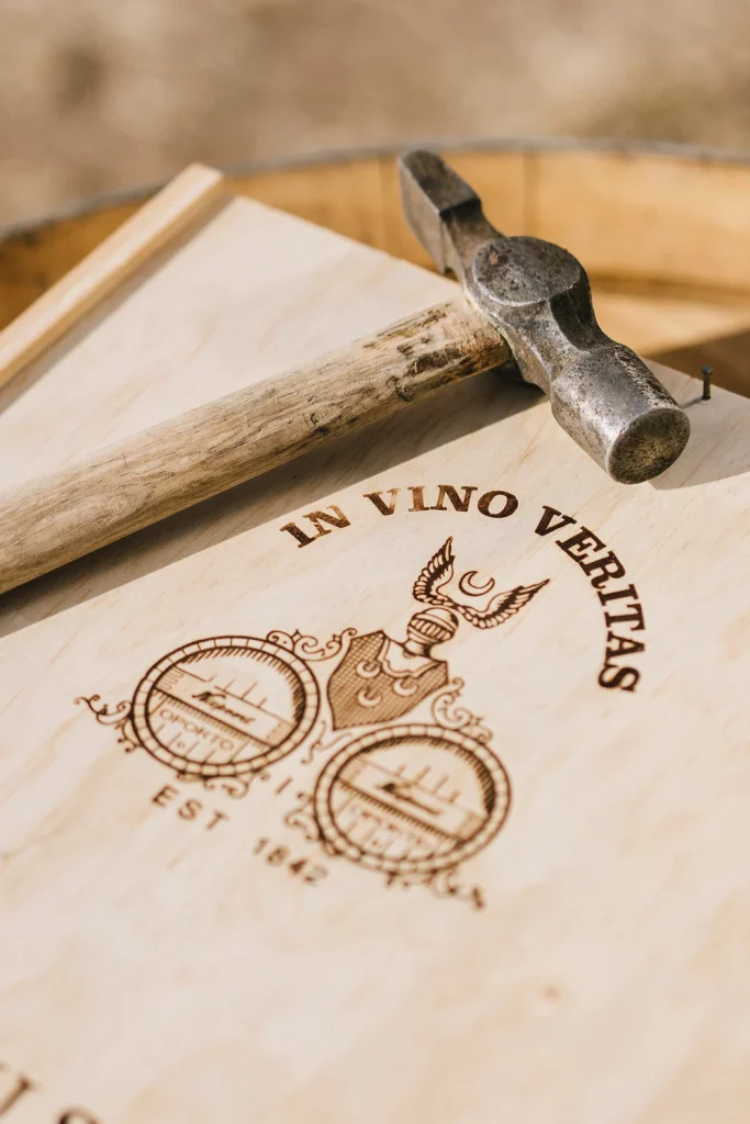 A close-up of a wooden wine box featuring an engraved crest and the phrase "In Vino Veritas," with a rustic hammer resting on top.
