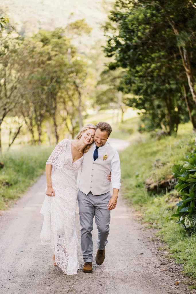 A bride and groom walk arm in arm along a scenic dirt path surrounded by greenery, laughing and sharing a joyful moment.
