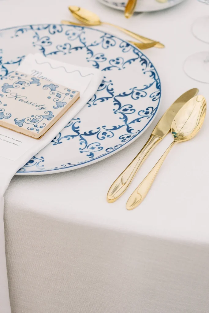 A wedding table setting featuring a blue and white patterned plate, gold cutlery, and a personalized ceramic name card.