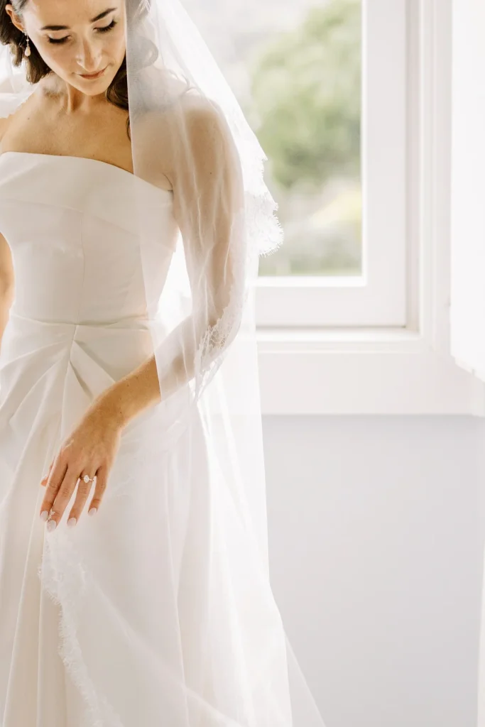 A bride gently adjusts her veil while standing by a window, bathed in soft natural light.