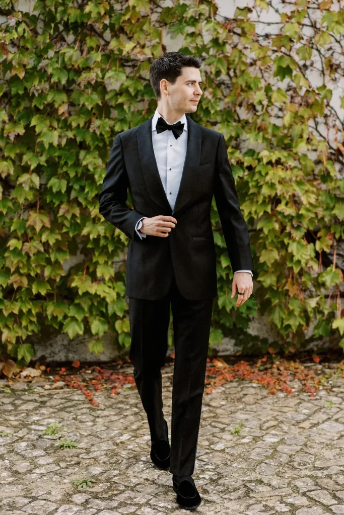 A groom in a classic black tuxedo walks confidently outside, with ivy-covered walls in the background.