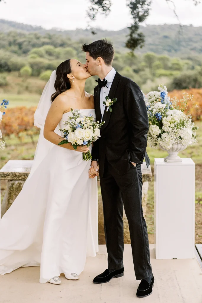 A groom stands with his back to the camera, his tuxedo jacket speckled with raindrops, as he faces his bride during their outdoor wedding ceremony.