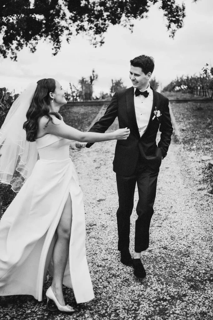 A black-and-white photo of a bride and groom walking hand in hand on a gravel path, smiling at each other in a vineyard setting.