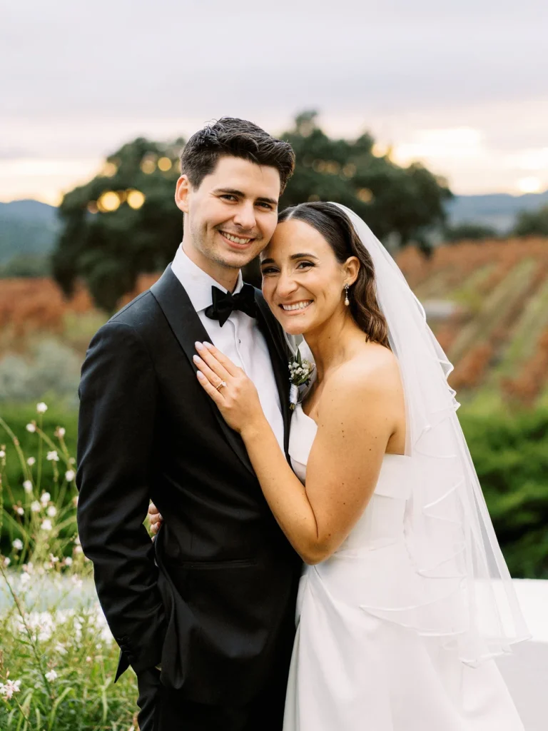 A newlywed couple poses together in a vineyard, smiling as they embrace, with a scenic backdrop of rolling hills and autumn vines.