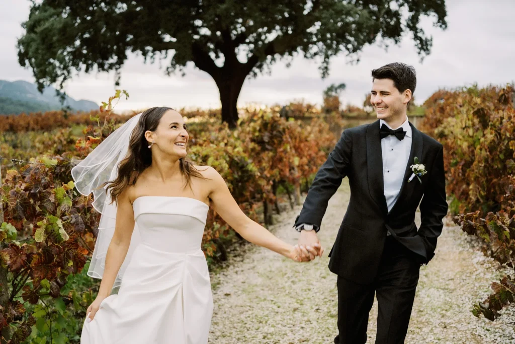 A newlywed couple walks hand in hand through a vineyard, smiling at each other with autumn foliage in the background.
