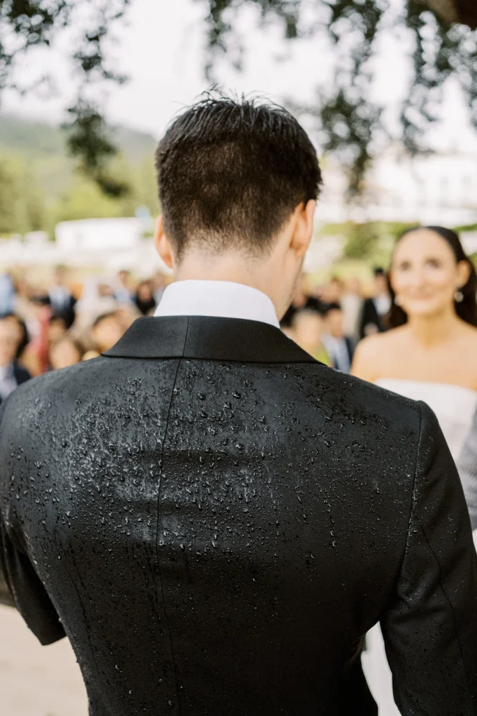 A newlywed couple walks up a stone path with wedding guests following behind through a scenic vineyard with rolling hills in the background.