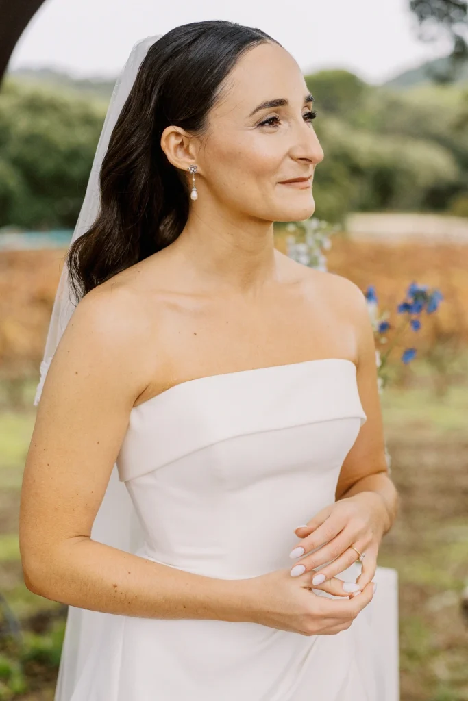 A bride and groom share a romantic kiss under a tree, surrounded by floral arrangements, with a vineyard landscape in the background.