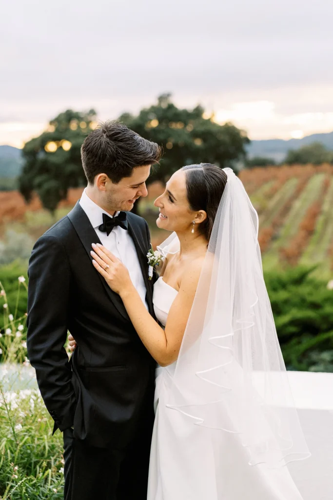 A bride and groom share a loving gaze in a vineyard at sunset, their faces close together with a backdrop of autumn vines.