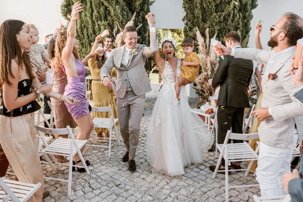 Newlyweds walk down the aisle with joyous expressions, holding hands as guests cheer and toss confetti at their outdoor wedding ceremony in Portugal. The bride carries a young child while guests celebrate around them.