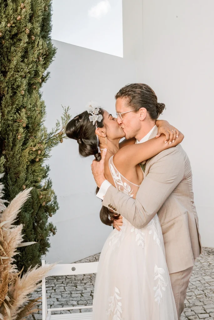 Newlyweds share a passionate kiss in front of a white chapel, surrounded by pampas grass and lush greenery.