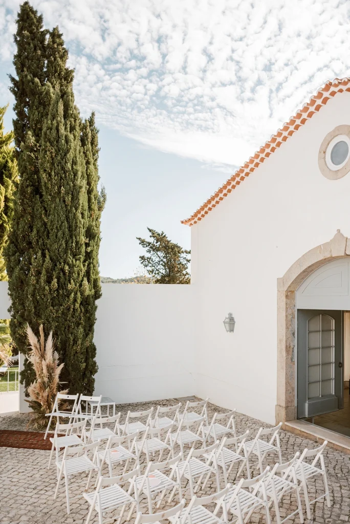 Elegant outdoor wedding ceremony space with white chairs arranged in front of a charming white chapel under a bright blue sky.