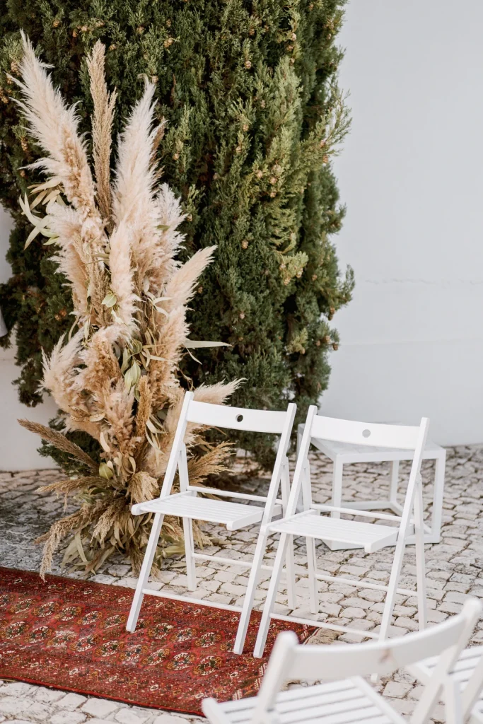 Outdoor wedding ceremony setup with white folding chairs, pampas grass arrangements, and a rustic rug in front of a tall cypress tree.
