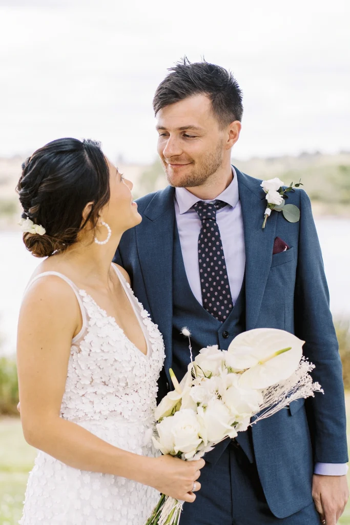 Bride and groom share a loving gaze outdoors, with the bride holding a bouquet of white flowers and the groom wearing a blue suit with a boutonniere.