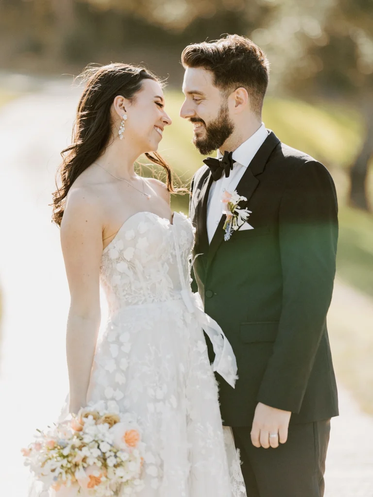 Bride and groom share a joyful gaze at sunset, with the bride in a lace gown holding a pastel bouquet and the groom in a black tuxedo with a boutonniere.