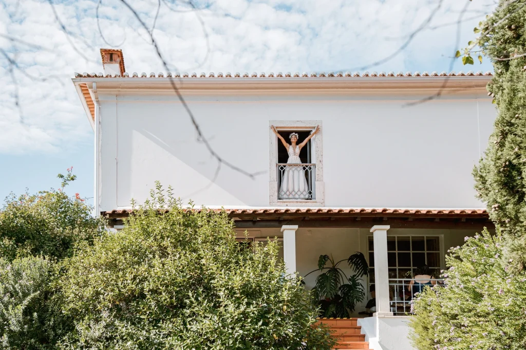 A joyful bride in a flowing wedding gown stands on a balcony with arms raised, framed by a white villa and lush greenery.