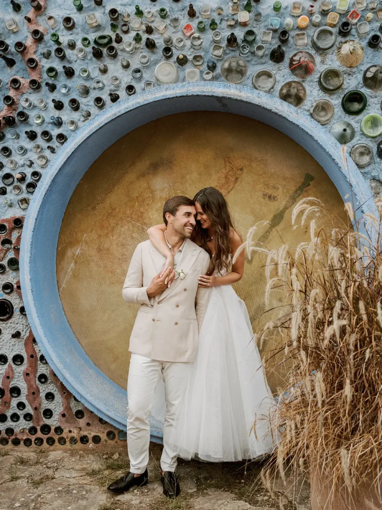 Bride and groom embracing in front of a vibrant circular bottle wall, sharing a joyful moment.