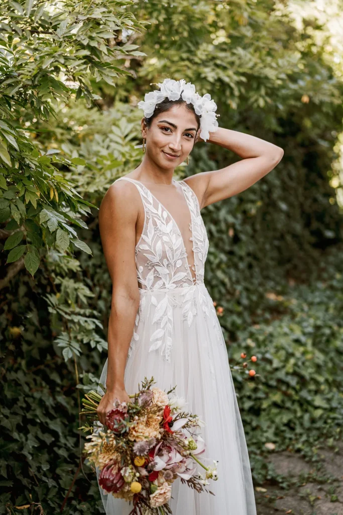 Bride smiling in a lush green garden, holding a colorful bouquet while adjusting her floral headpiece.